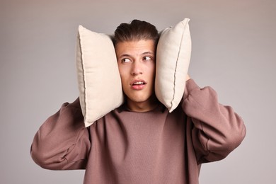 Photo of Annoyed young man covering his ears with pillows due to loud sound on light grey background