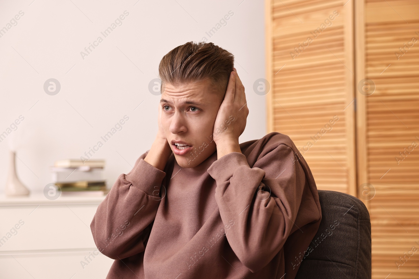 Photo of Annoyed young man covering his ears due to loud sound at home