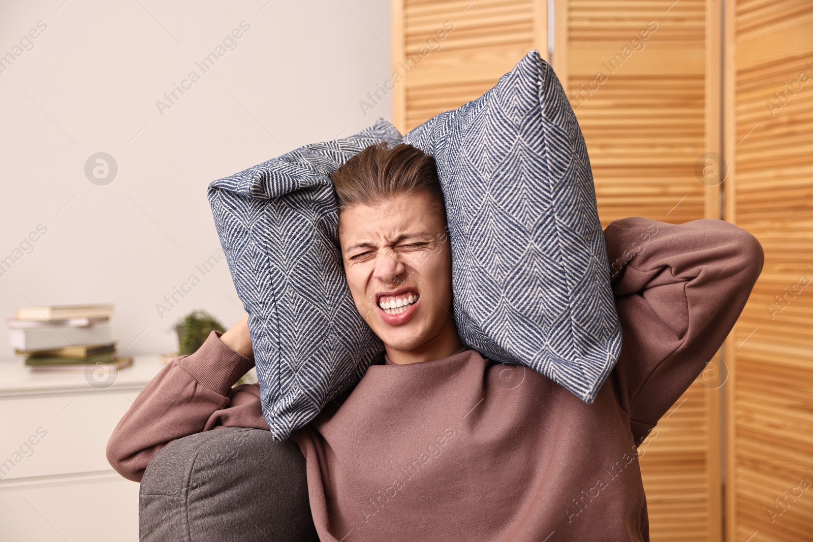 Photo of Annoyed young man covering his ears with pillows due to loud sound at home