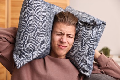 Photo of Annoyed young man covering his ears with pillows due to loud sound at home