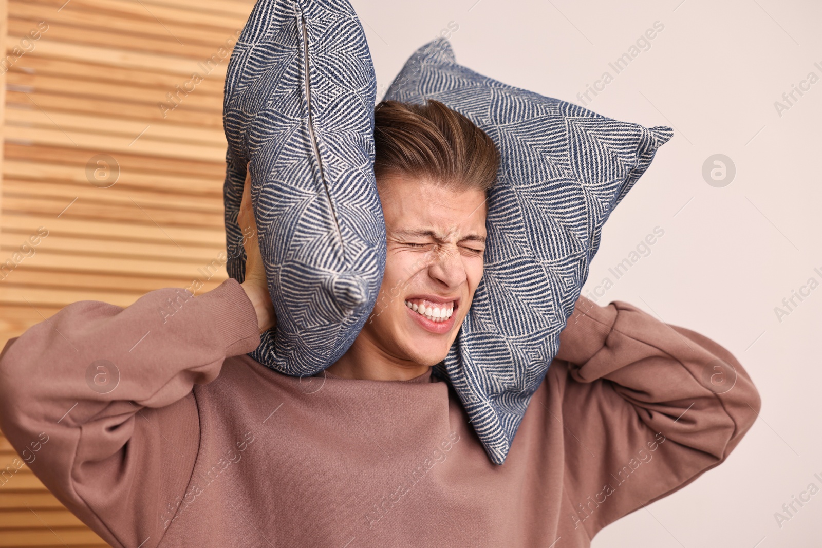 Photo of Annoyed young man covering his ears with pillows due to loud sound at home