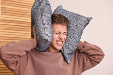 Photo of Annoyed young man covering his ears with pillows due to loud sound at home