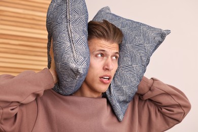 Photo of Annoyed young man covering his ears with pillows due to loud sound at home