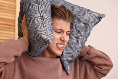 Photo of Annoyed young man covering his ears with pillows due to loud sound at home