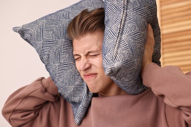 Photo of Annoyed young man covering his ears with pillows due to loud sound at home