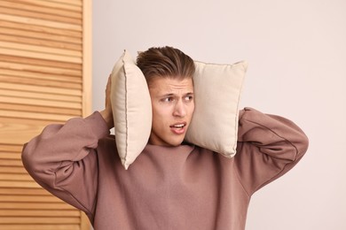 Photo of Annoyed young man covering his ears with pillows due to loud sound at home