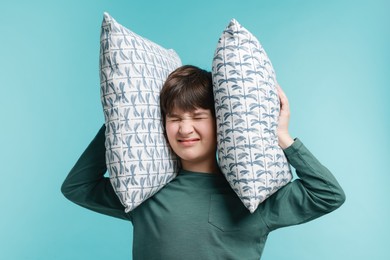 Photo of Annoyed boy covering his ears with pillows due to loud sound on light blue background