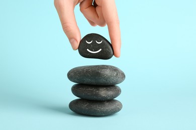 Photo of Woman putting rock with smiley face onto cairn against light blue background, closeup. Harmony and life balance