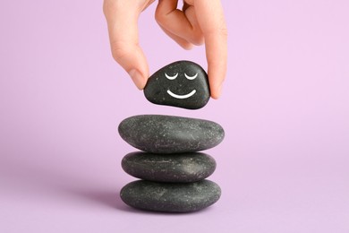 Photo of Woman putting rock with smiley face onto cairn against lilac background, closeup. Harmony and life balance