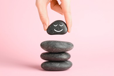 Photo of Woman putting rock with smiley face onto cairn against light pink background, closeup. Harmony and life balance