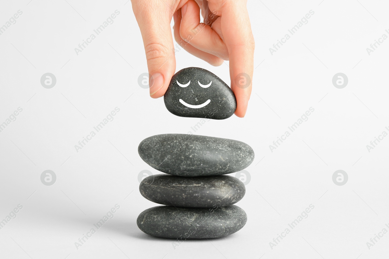 Photo of Woman putting rock with smiley face onto cairn against light background, closeup. Harmony and life balance