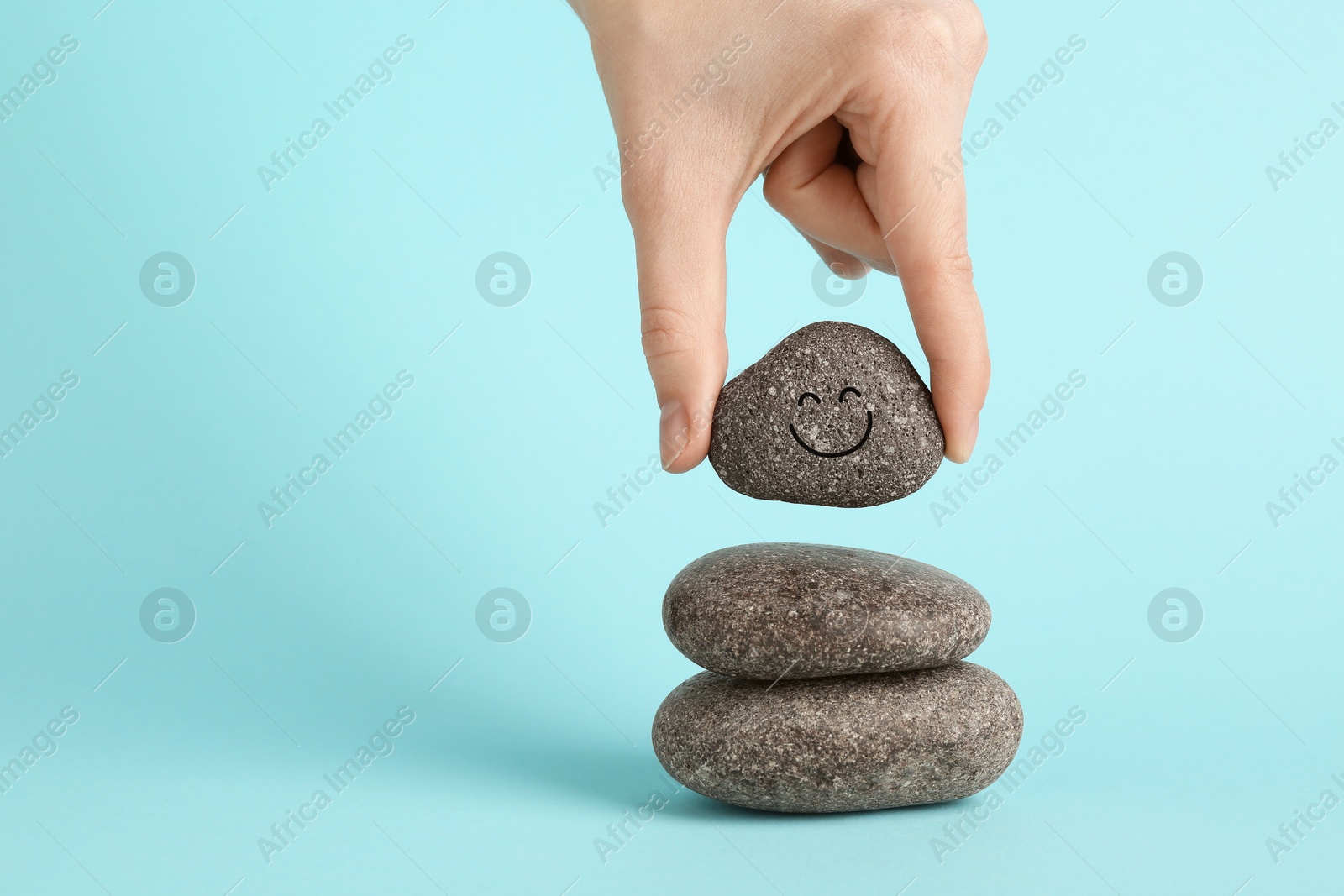 Photo of Woman putting rock with smiley face onto cairn against light blue background, closeup and space for text. Harmony and life balance