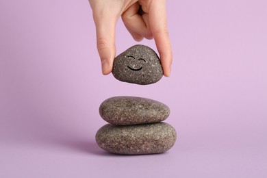 Photo of Woman putting rock with smiley face onto cairn against lilac background, closeup. Harmony and life balance