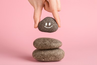 Photo of Woman putting rock with smiley face onto cairn against light pink background, closeup. Harmony and life balance