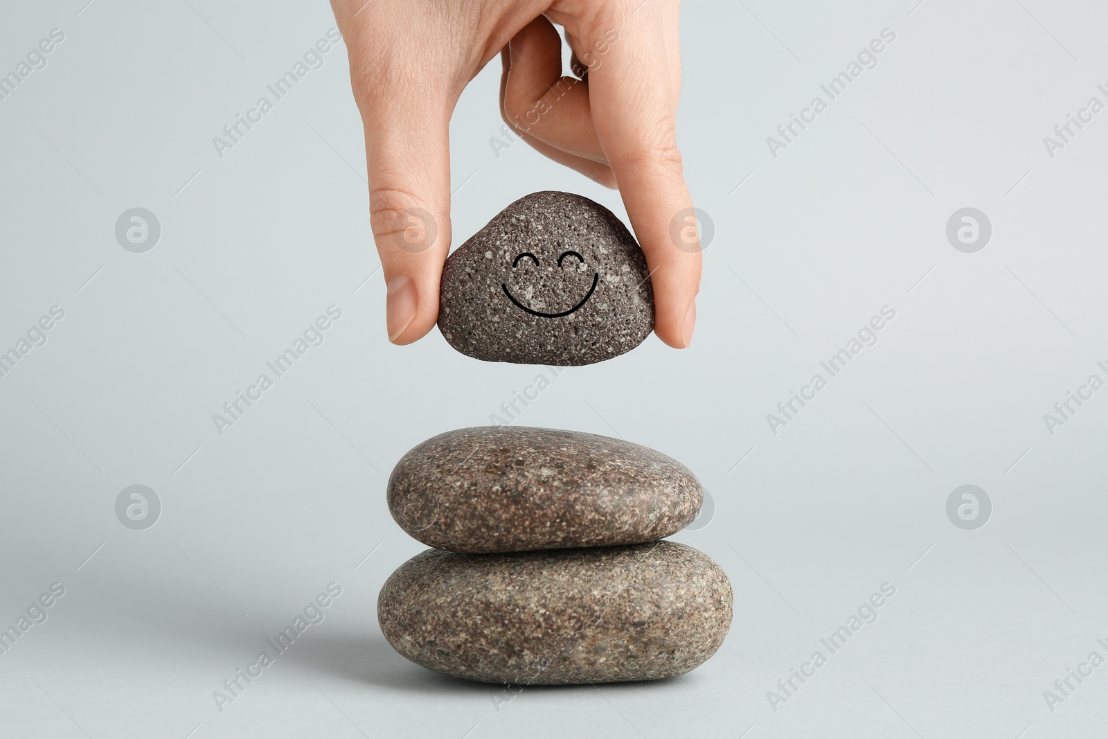 Photo of Woman putting rock with smiley face onto cairn against light grey background, closeup. Harmony and life balance