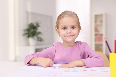 Photo of Cute girl at white table with drawing and pencils in kindergarten