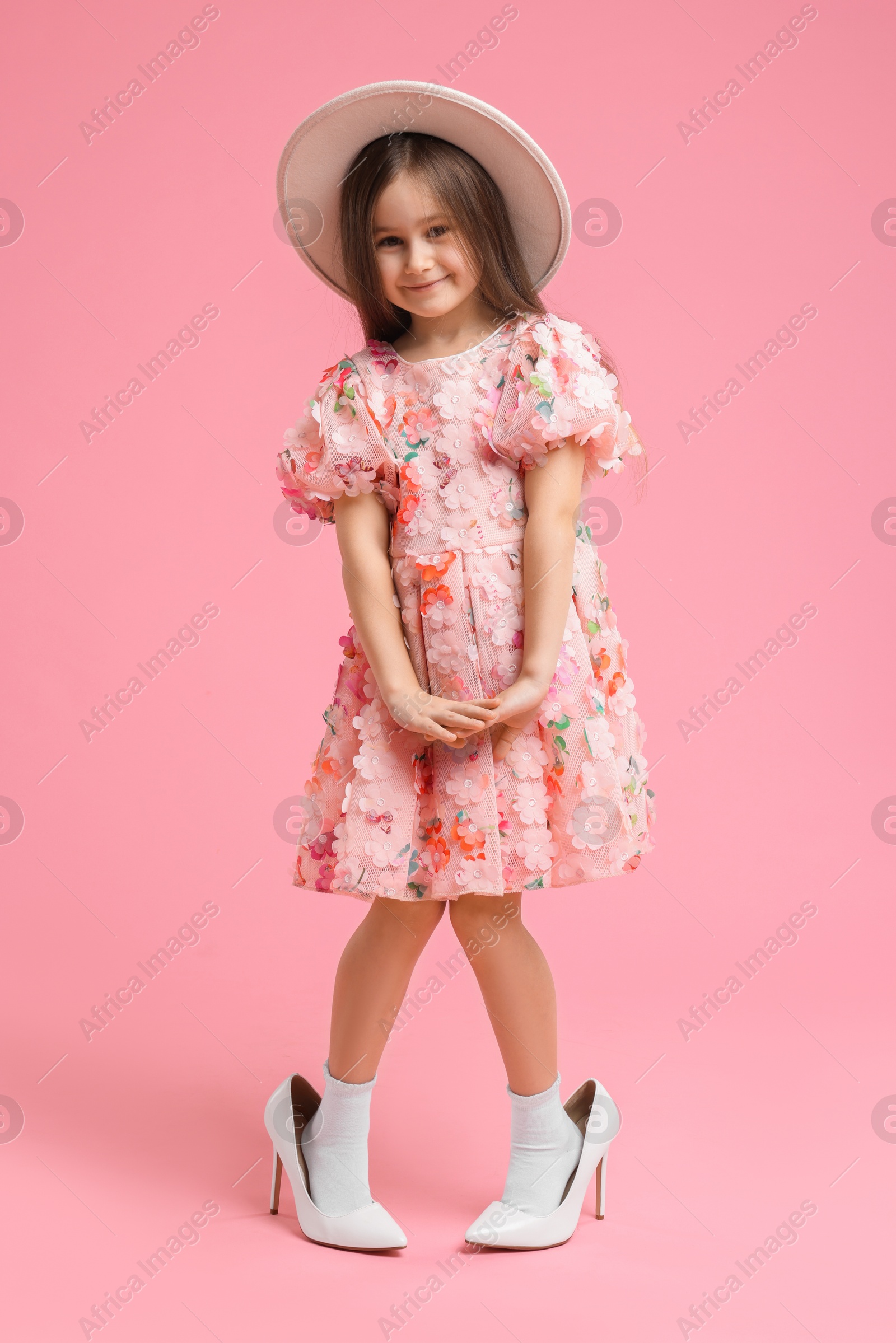 Photo of Stylish little girl wearing oversized high heeled shoes on pink background