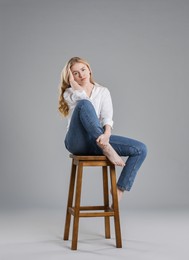 Photo of Beautiful young woman in stylish jeans sitting on stool against grey background