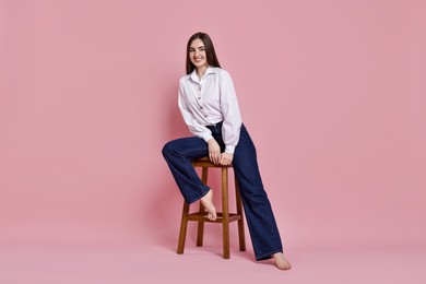 Photo of Smiling woman in stylish jeans sitting on stool against pink background