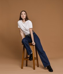 Photo of Beautiful young woman in stylish jeans sitting on stool against brown background