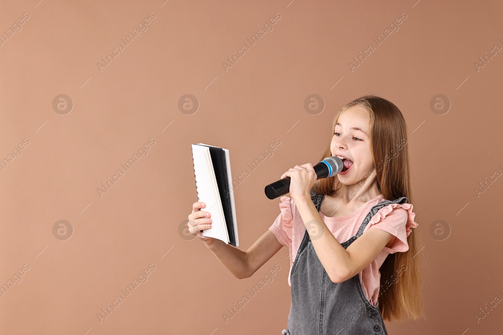 Photo of Little girl with microphone and notebook singing on light brown background, space for text