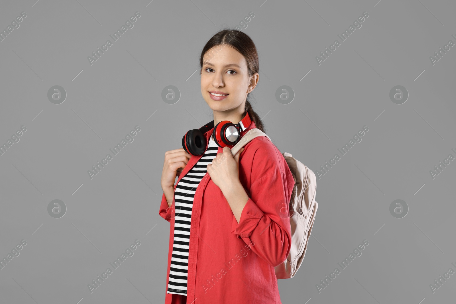 Photo of Portrait of smiling teenage girl with backpack and headphones on grey background