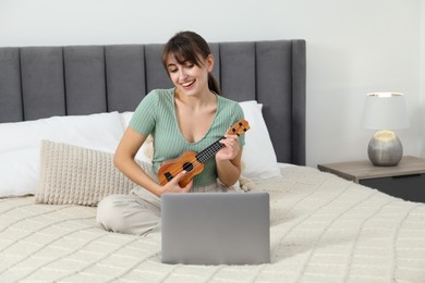 Photo of Happy woman learning to play ukulele with online music course on bed at home