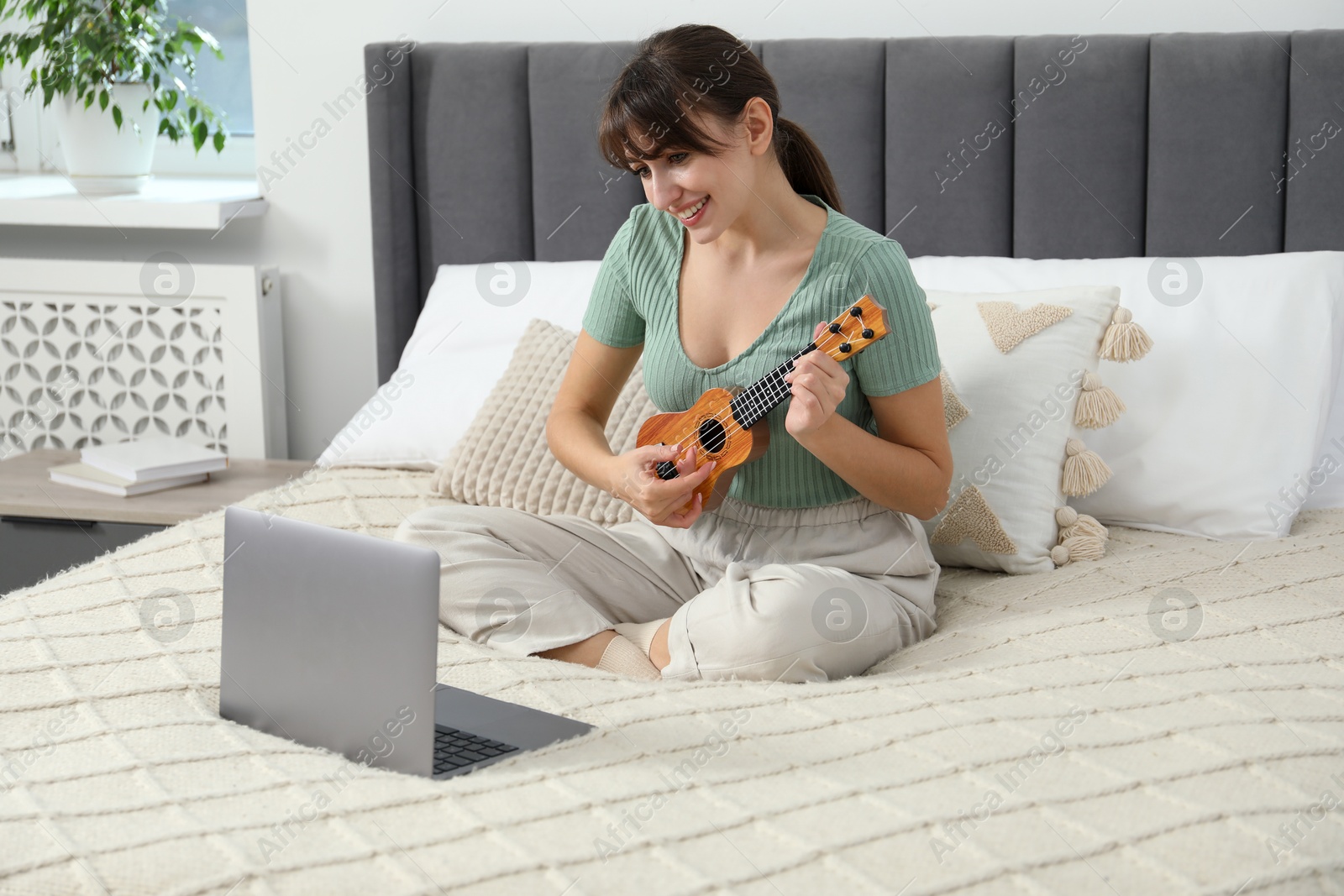 Photo of Happy woman learning to play ukulele with online music course on bed at home