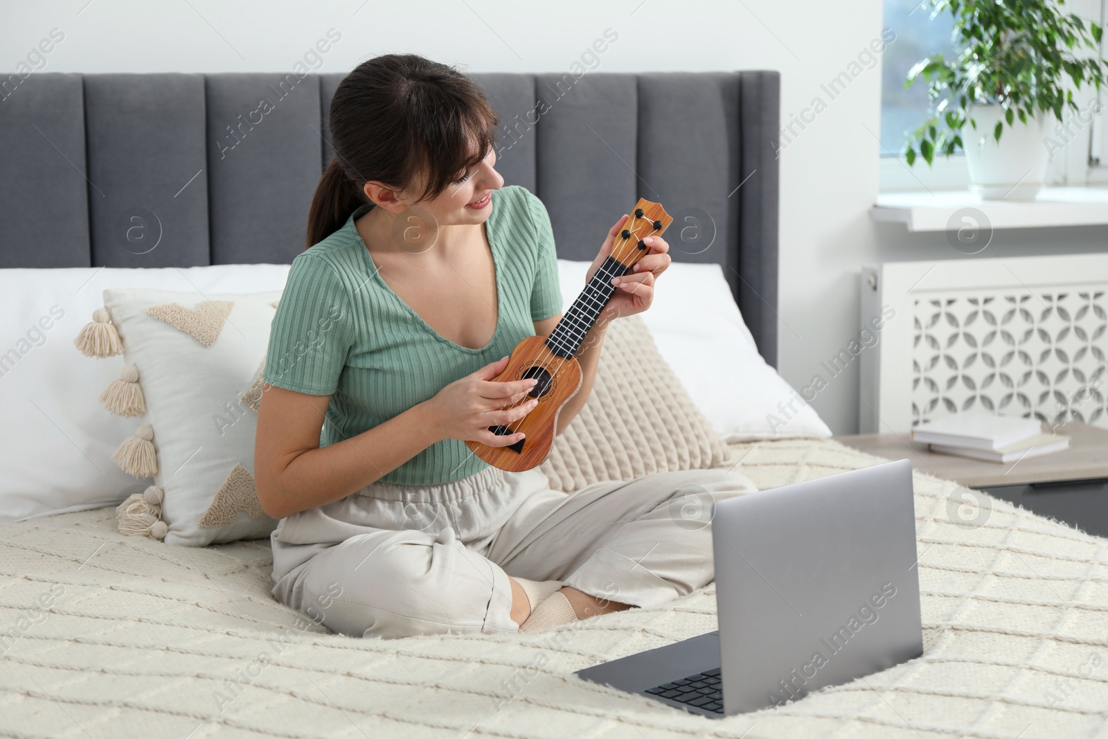 Photo of Happy woman learning to play ukulele with online music course on bed at home