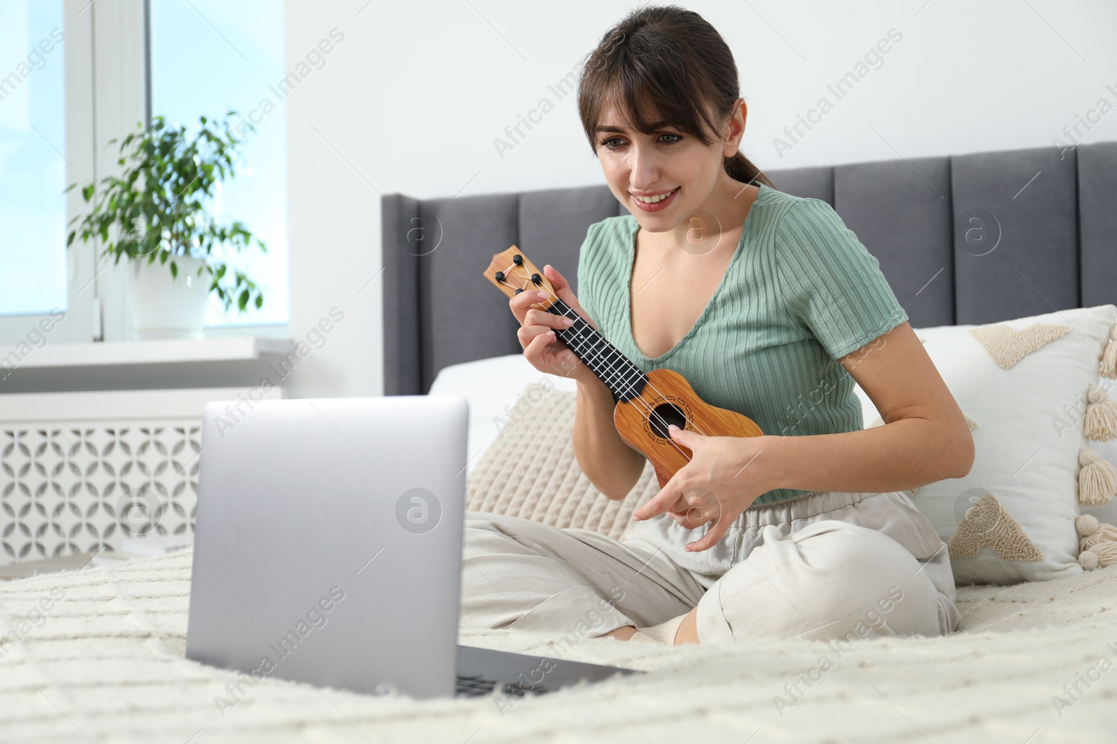 Photo of Happy woman learning to play ukulele with online music course on bed at home