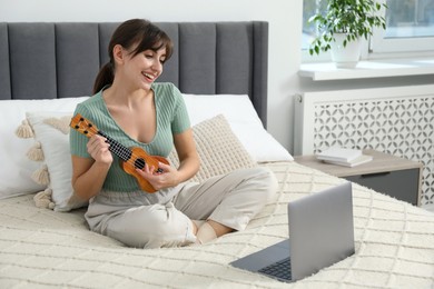 Photo of Happy woman learning to play ukulele with online music course on bed at home