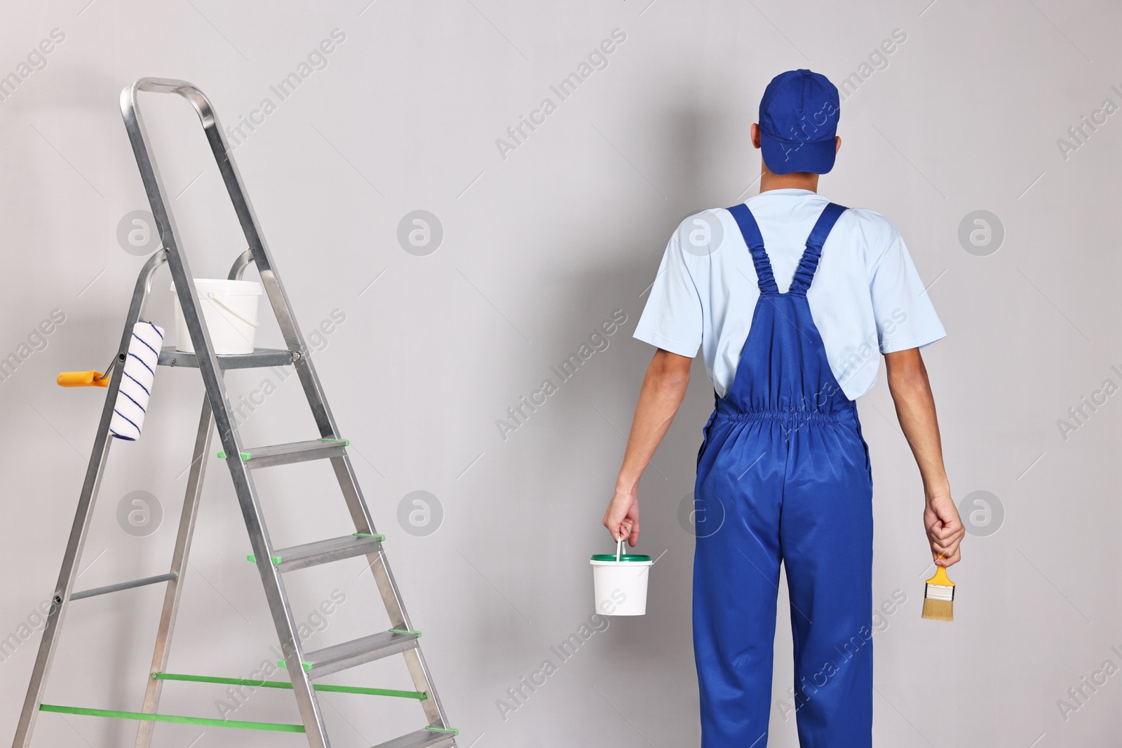 Photo of Male handyman with brush and bucket of paint near ladder indoors, back view