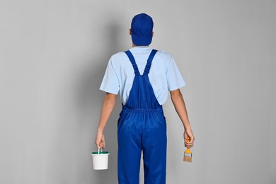 Photo of Male handyman with brush and bucket of paint near grey wall, back view