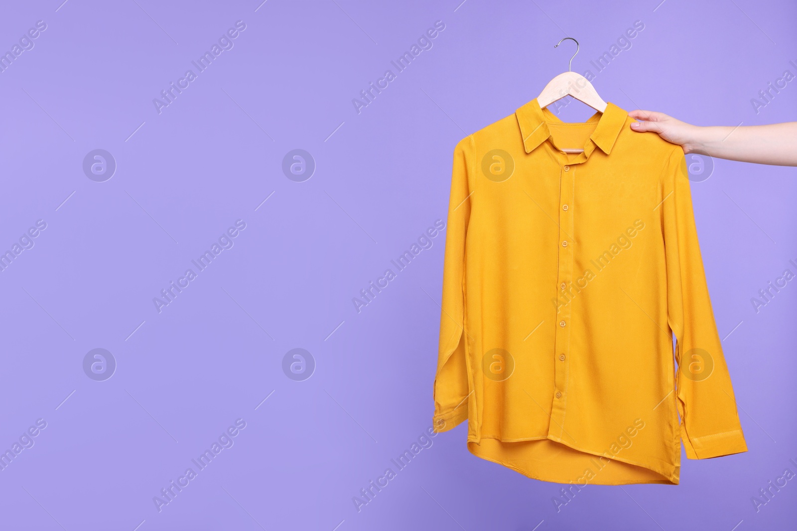 Photo of Woman holding hanger with orange shirt on light purple background, closeup. Space for text