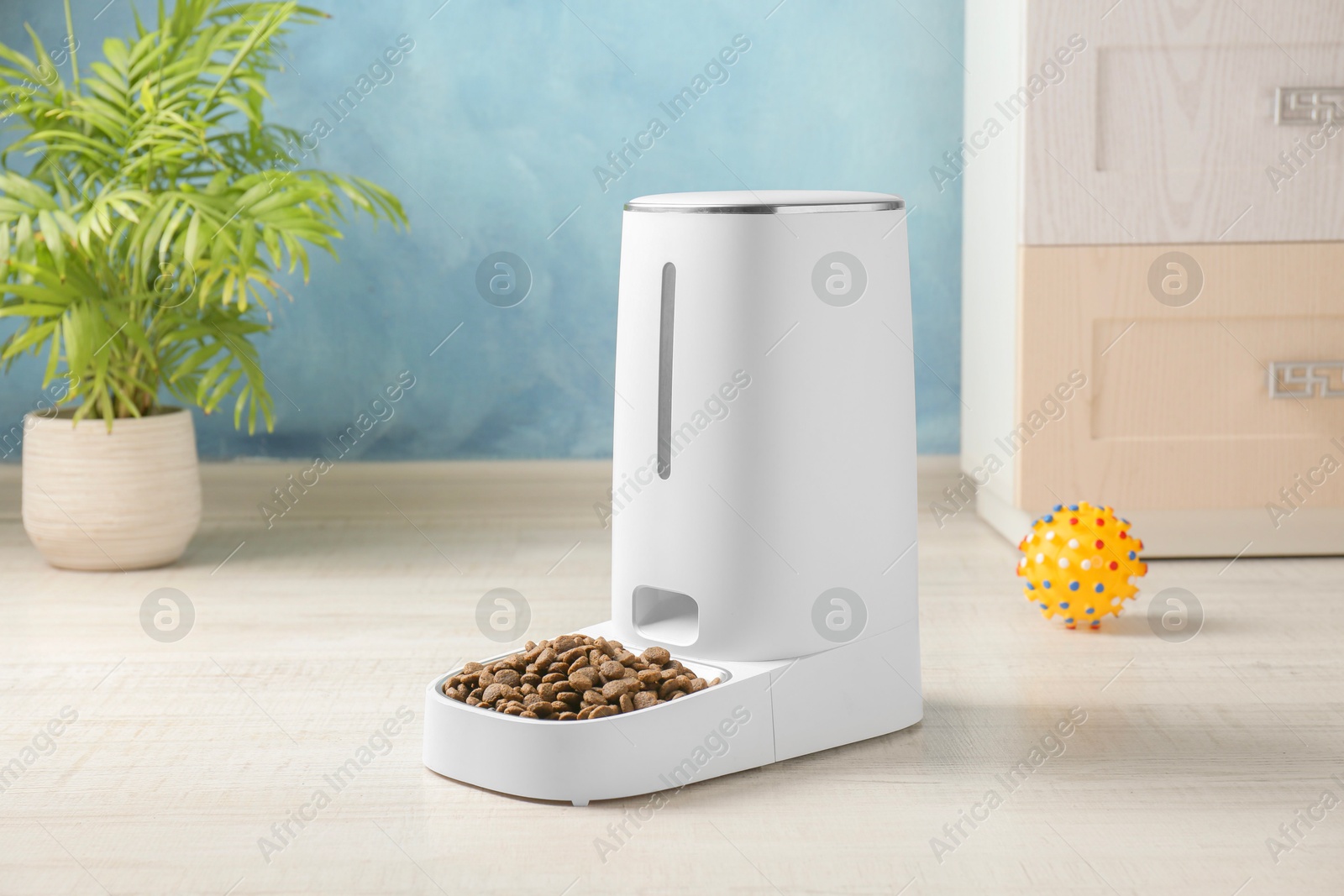 Photo of Dry pet food in automatic feeder and toy on floor indoors