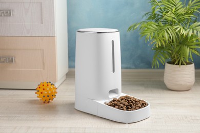 Photo of Dry pet food in automatic feeder and toy on floor indoors