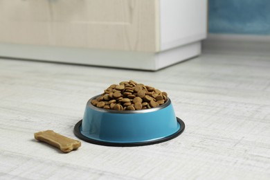 Photo of Dry pet food in feeding bowl and chew bone on floor indoors