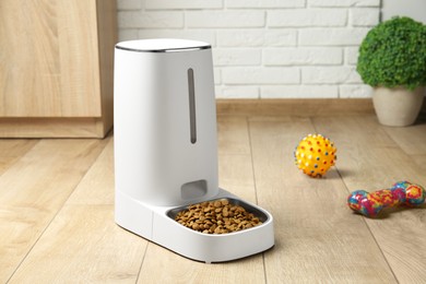 Photo of Dry pet food in automatic feeder and toys on floor indoors