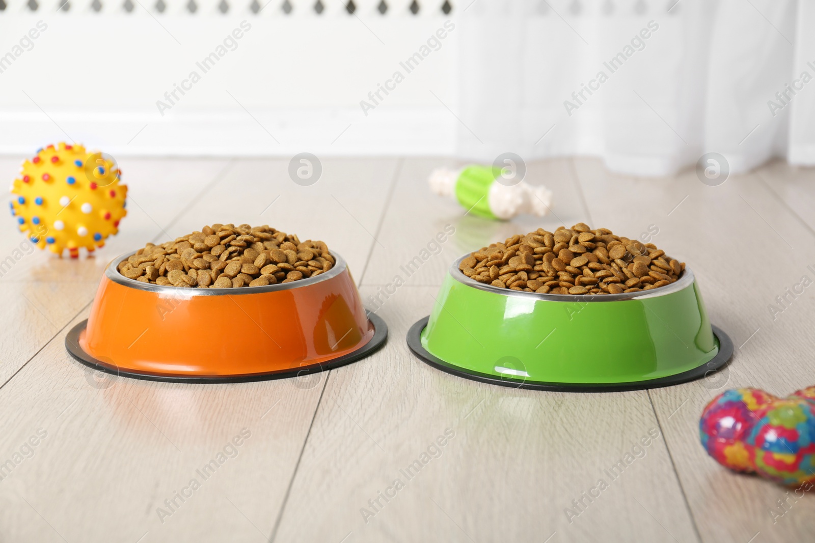 Photo of Dry pet food in feeding bowls and toys on floor indoors