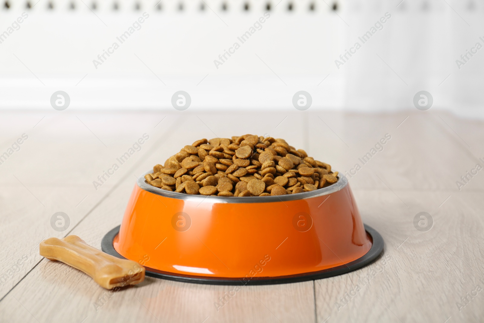 Photo of Dry pet food in feeding bowl and chew bone on floor indoors, closeup