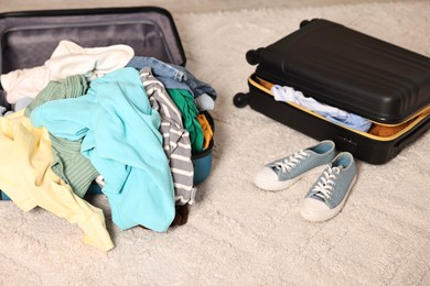 Photo of Suitcase with pile of different messy clothes, another one and sneakers on carpet
