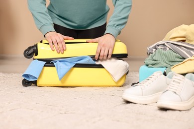 Photo of Woman trying to close suitcase full of different messy clothes indoors, closeup