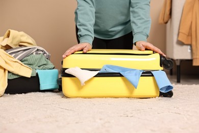 Photo of Woman trying to close suitcase full of different messy clothes indoors, closeup