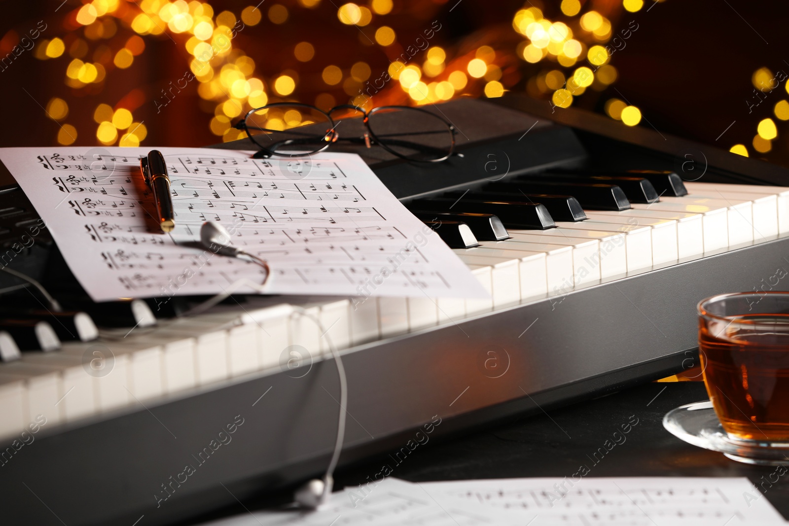 Photo of Piano, musical notes, pen and cup of tea on table against blurred lights, closeup. Bokeh effect