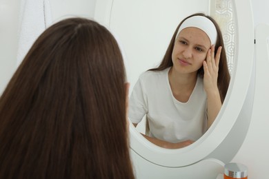 Photo of Upset teenage girl with acne problem looking in mirror indoors