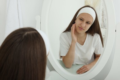 Photo of Upset teenage girl with acne problem looking in mirror indoors