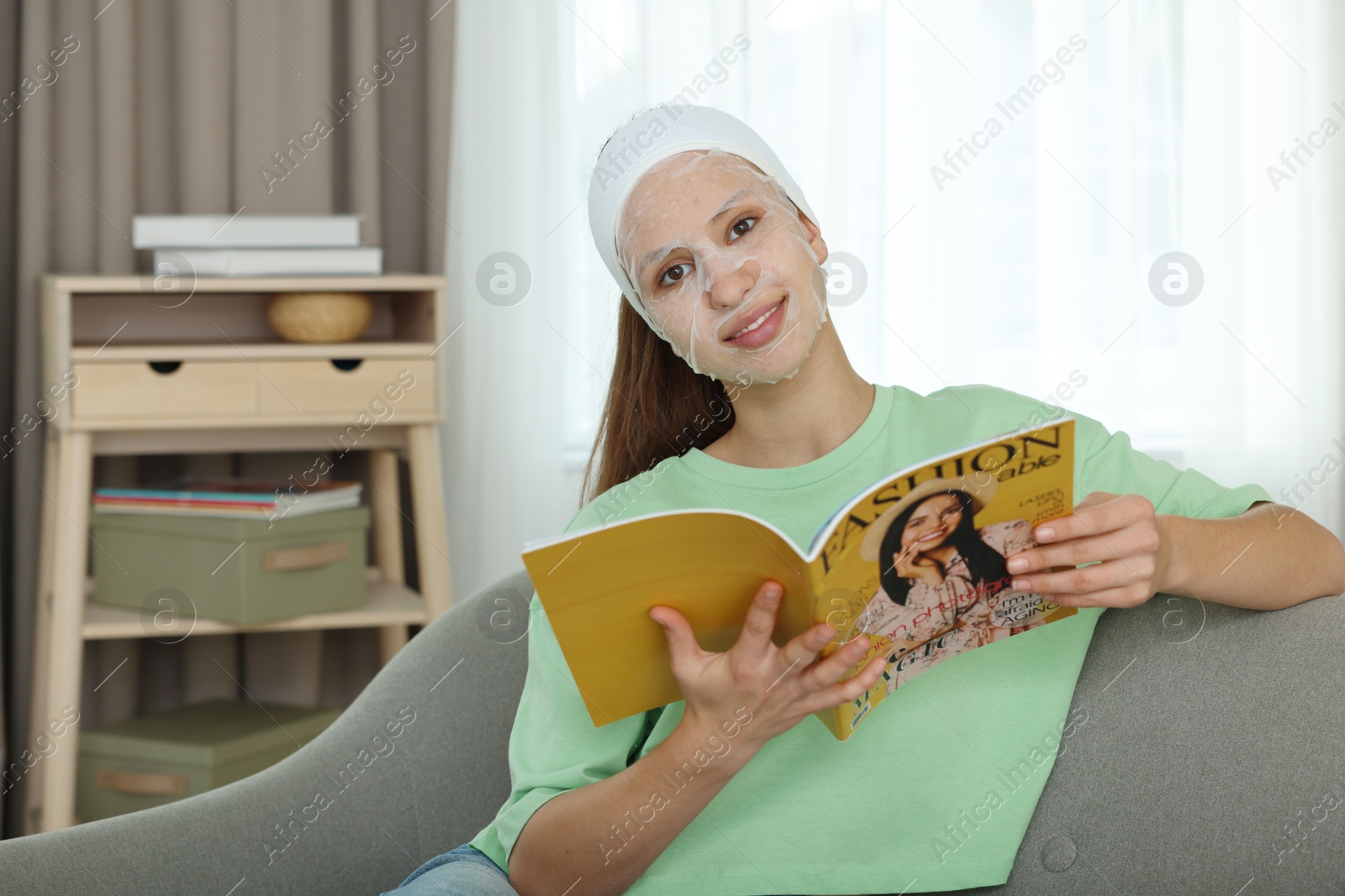 Photo of Teenage girl with sheet facial mask reading magazine at home. Acne treatment