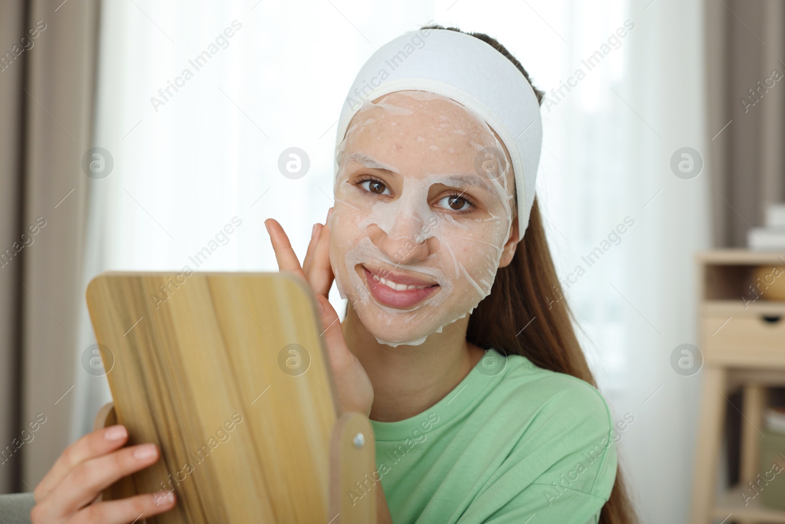 Photo of Teenage girl with sheet facial mask looking in mirror indoors. Acne treatment