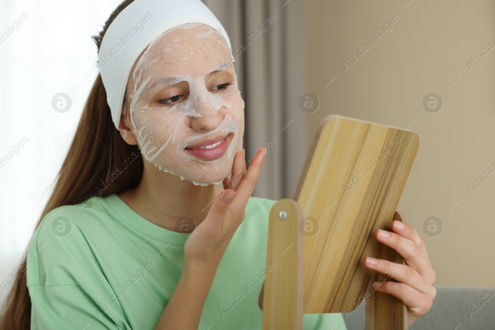 Photo of Teenage girl with sheet facial mask looking in mirror indoors. Acne treatment