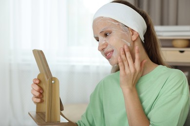 Photo of Teenage girl with sheet facial mask looking in mirror indoors. Acne treatment